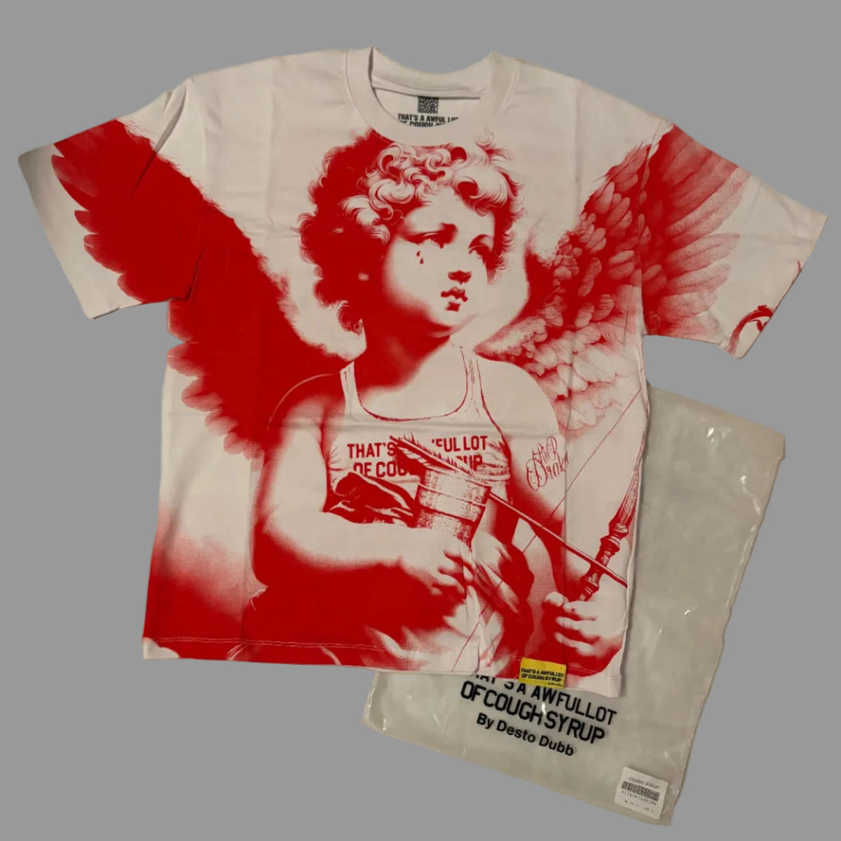 A white graphic t-shirt featuring a red and white print of a winged cherub holding a syringe, laid flat on a wrinkled white sheet next to a clear plastic bag with printed text.
