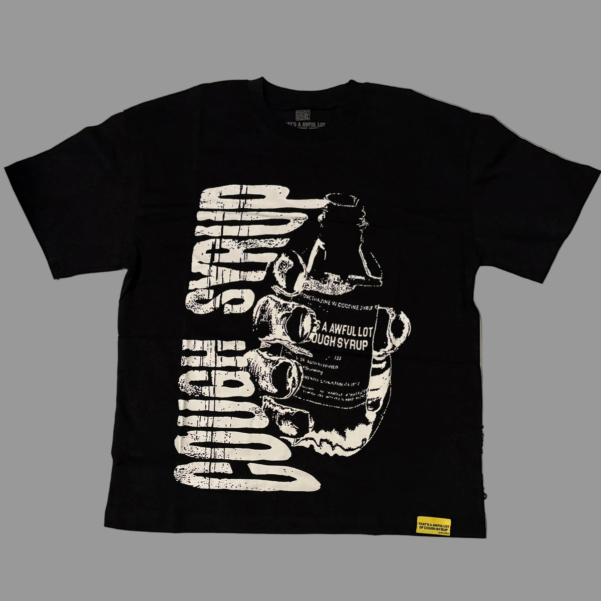 A black short-sleeve graphic t-shirt laid flat on a white wrinkled surface, featuring a large distressed white print of a cough syrup bottle and the words 'COUGH SYRUP' and '2 A HUNTER LOT COUGH SYRUP'. A clear plastic bag with similar text is partially visible on the right.
