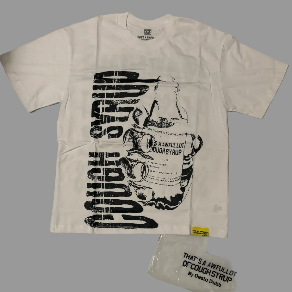 A white short-sleeve graphic T-shirt laid flat on a light background, featuring a large black distressed print with the words 'Cough Syrup' and an illustration of a bottle. A clear plastic bag with matching printed text is placed beside it.