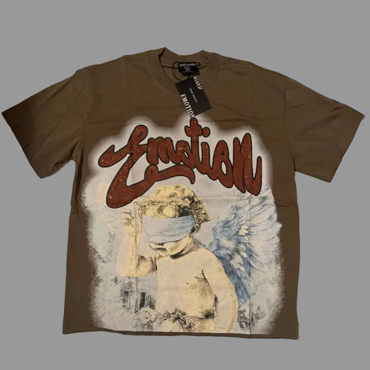 A brown short-sleeve graphic t-shirt laid flat on a white fabric background. The shirt features a large front print with the word 'Emotion' in red graffiti-style lettering above an illustration of a blindfolded cherub with blue wings.